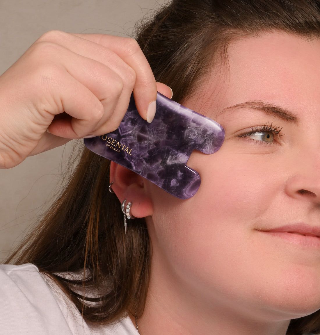Frau verwendet Amethyst Gua Sha. Lila Stein wird am Gesicht gehalten. Logo sichtbar.