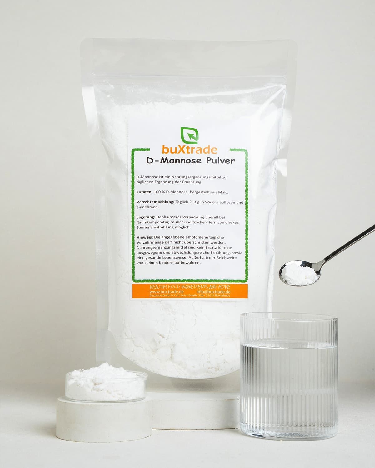 Ein Beutel D-Mannose Pulver von buxtrade neben einem Glas Wasser, einem Löffel mit Pulver und einer kleinen Schale.