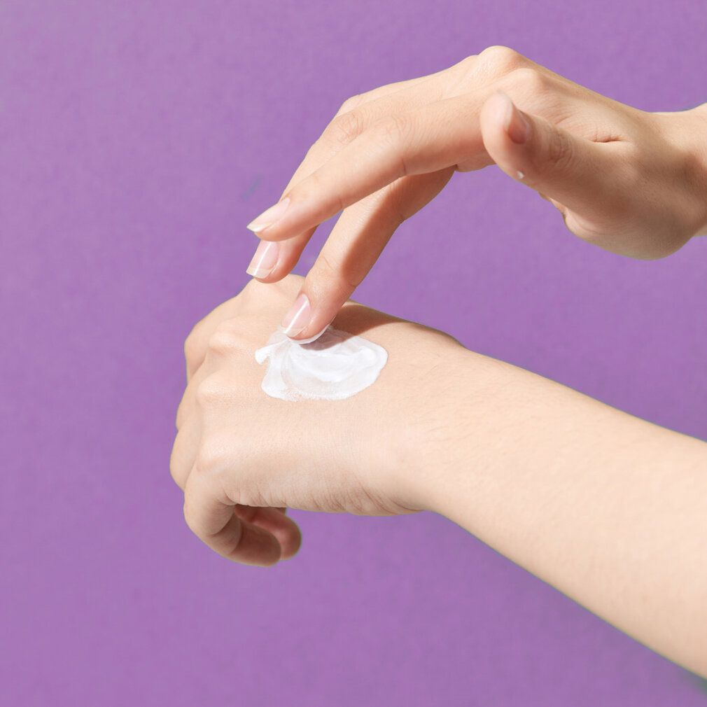 Hände tragen Creme auf. Weißliche Creme auf lila Hintergrund.