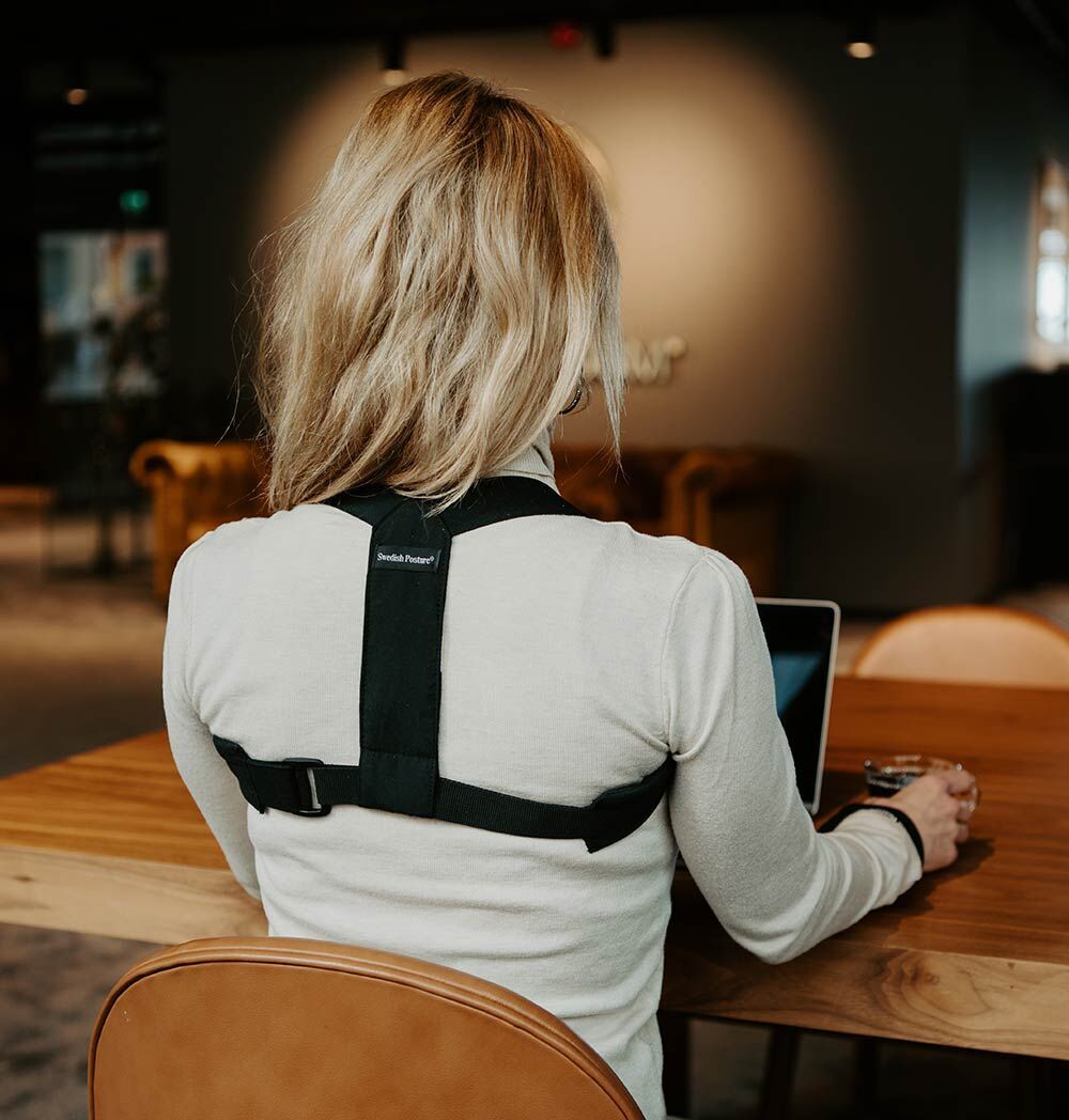 Eine Frau sitzt an einem Tisch und arbeitet an einem Laptop. Sie trägt eine schwarze Haltungsstütze unter ihrer Kleidung.