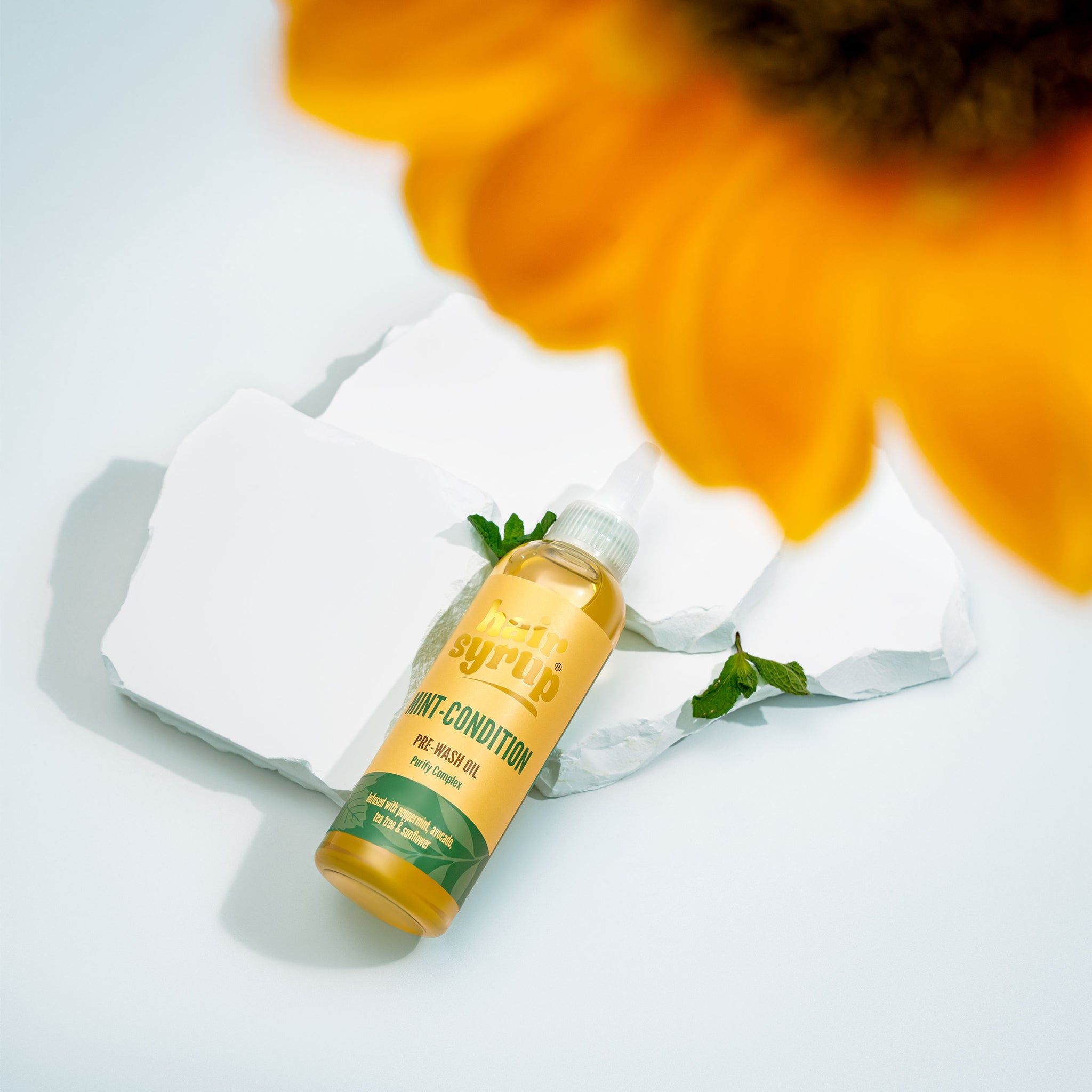 Gelbe Flasche mit Aufschrift "Hair Syrup Mint Condition". Auf weißen Steinen platziert, mit Minze und Blume.