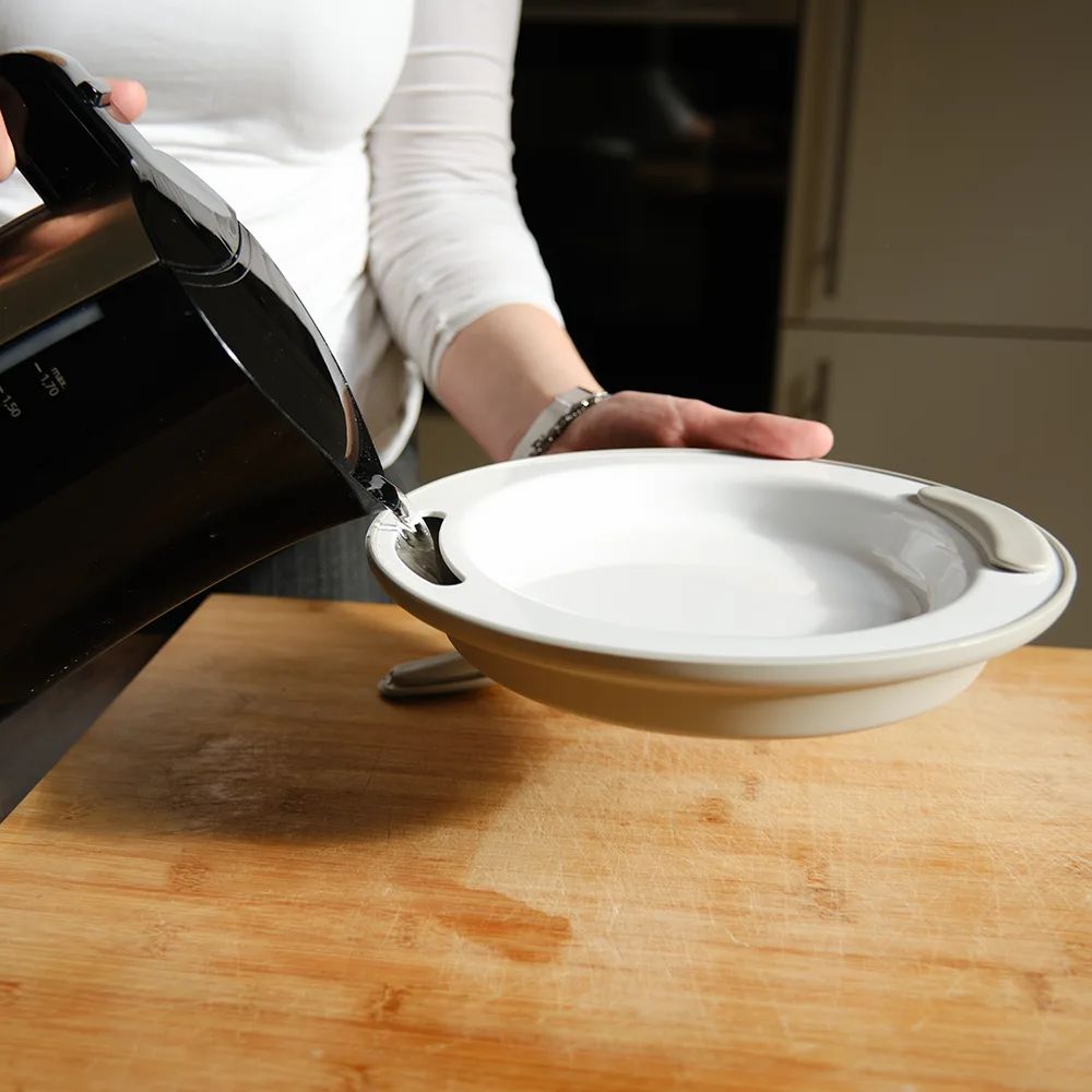 Person gießt Wasser in einen Teller mit Griffen. Der Teller steht auf einem Holztisch.