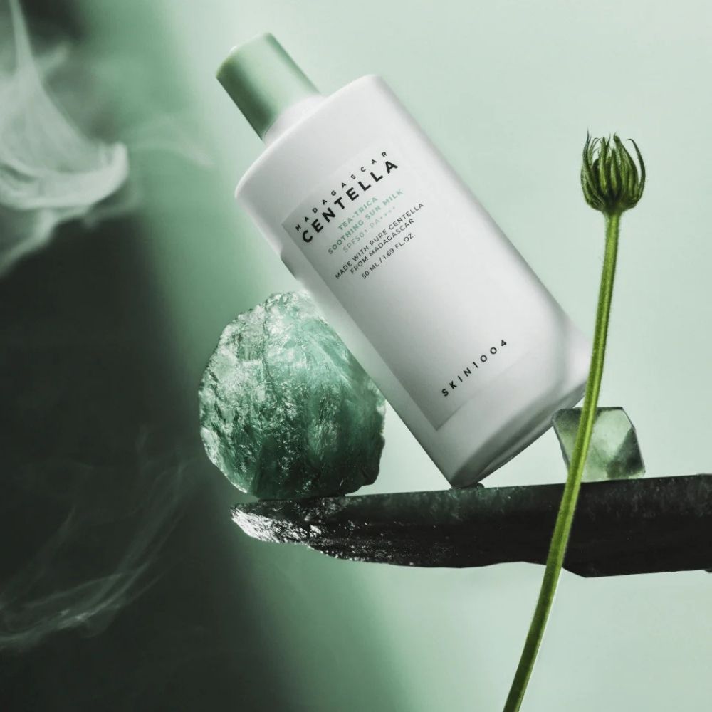 Flasche Madagascar Centella Tea-Trica Soothing Sun Milk SPF50+ PA++++ auf Stein mit grünen Kristallen und Rauch.