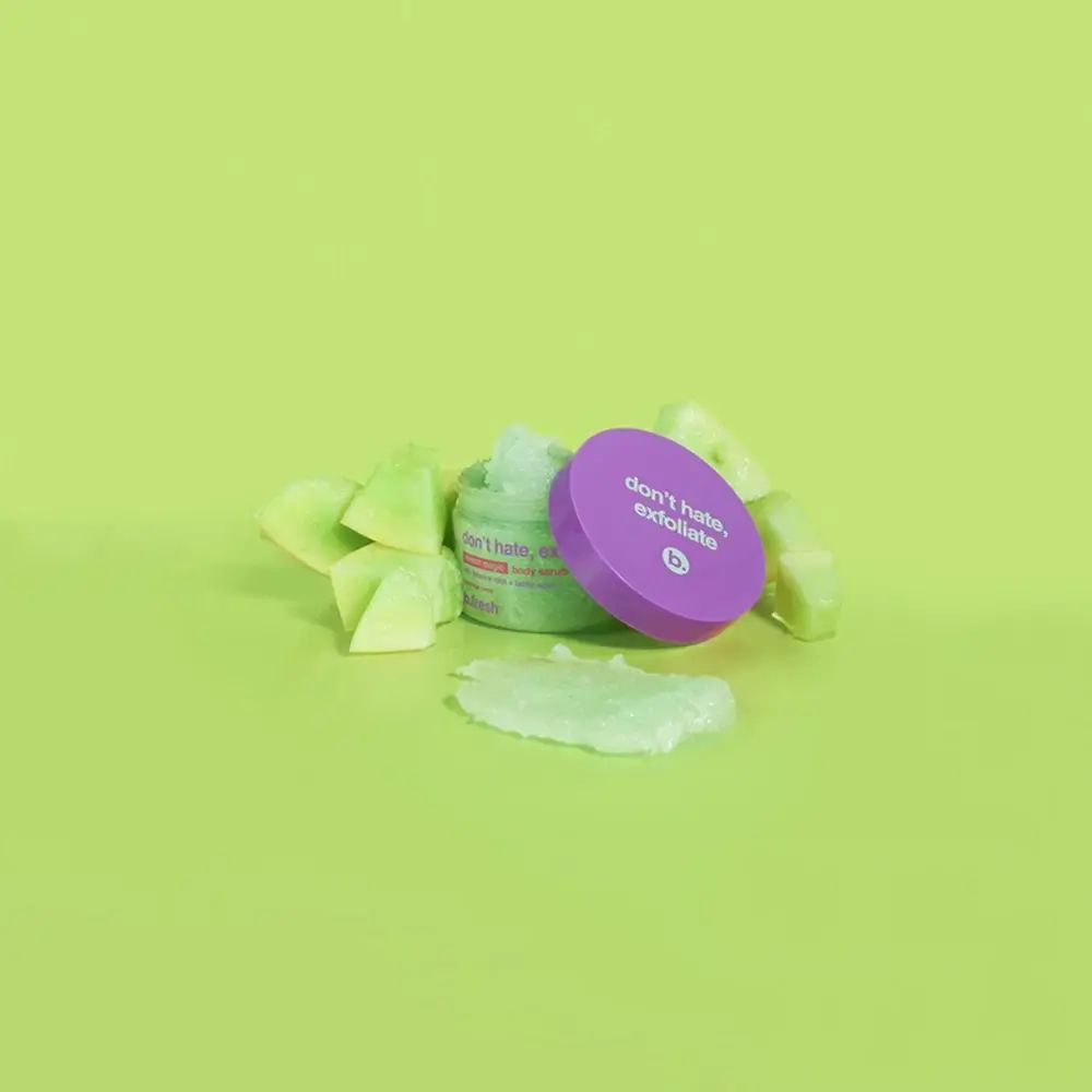 Offener Behälter mit Körperpeeling und Deckel neben Melonenstücken auf grünem Hintergrund.