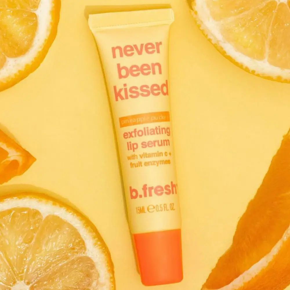 Gelbe Tube mit rotem Verschluss, umgeben von Zitronen. Aufschrift: "never been kissed exfoliating lip serum" und "b.fresh".