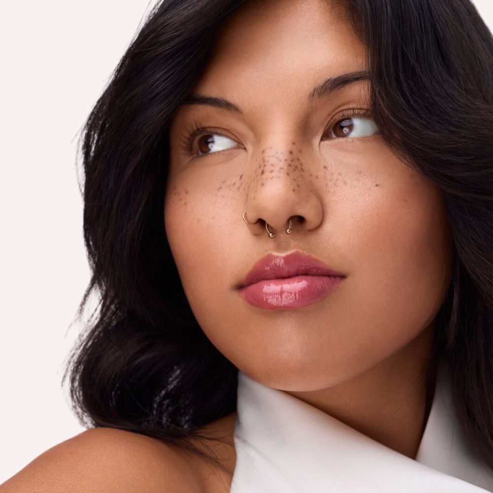 Porträt einer Frau mit aufgetragenem rosa Lippenfiller. Dunkles Haar, Sommersprossen, Nasenpiercing.