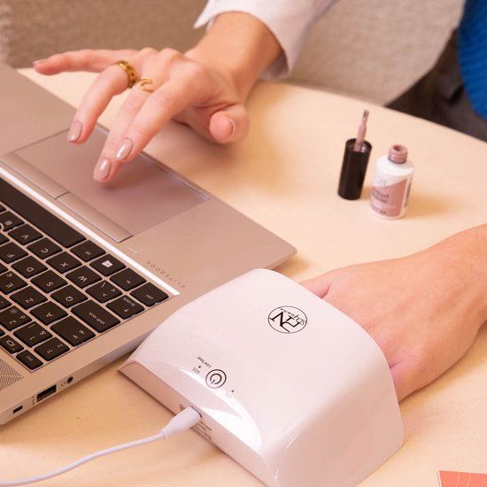 Person arbeitet am Laptop. UV-Lampe und Nagellackflasche auf dem Tisch. Lampe weiß, mit Logo.
