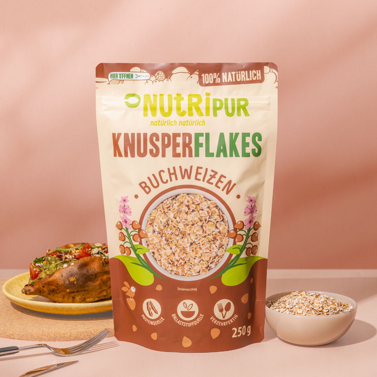 Verpackung NutriPur Knusperflakes Buchweizen. Beutel mit Flocken, 250g. Auf Teller: Ofenkartoffel mit Belag. Gabeln.