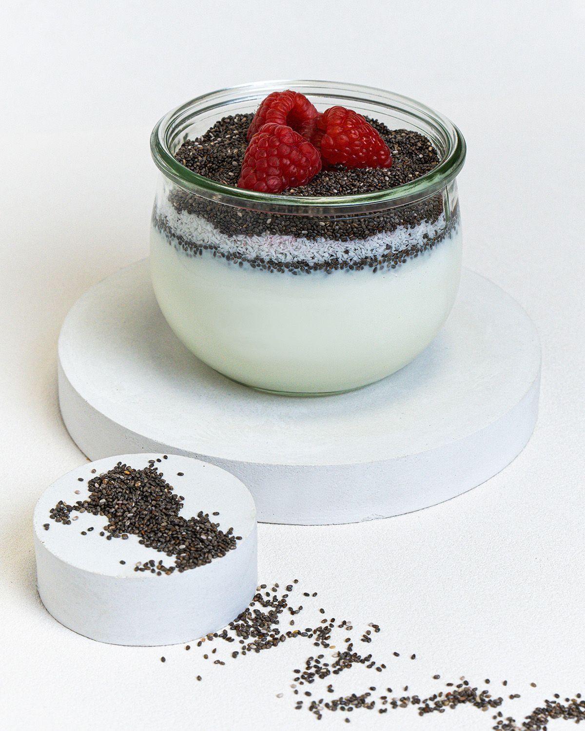 Glas mit Chia-Samen, Joghurt und Himbeeren. Auf weißem Podest. Streuung von Samen.
