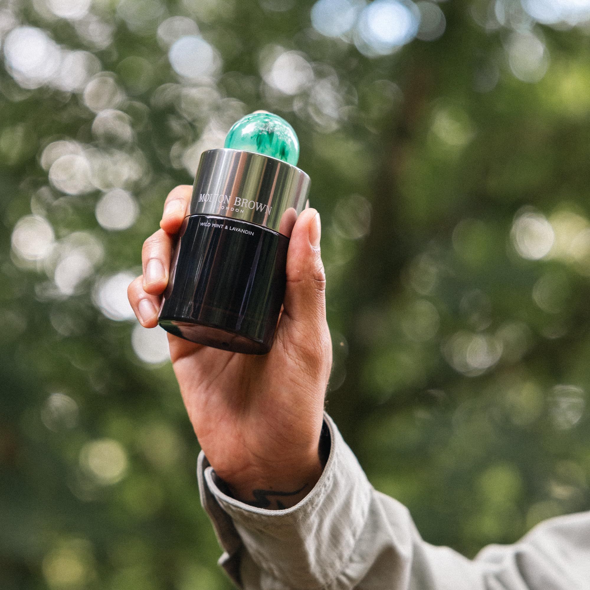 Hand hält eine dunkle Glasflasche mit grünem Deckel. Aufschrift: Wild Mint & Lavandin. Hintergrund: verschwommener Wald.