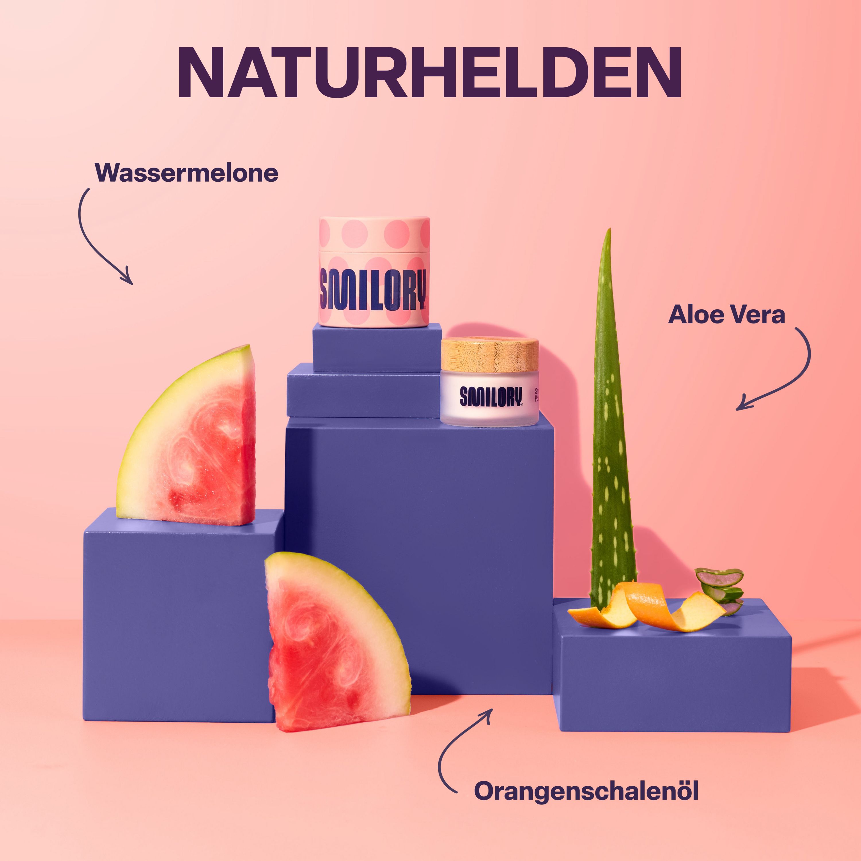 Produkt-Arrangement mit Wassermelone, Aloe Vera und Orangenöl. Zwei Smilory-Behälter auf blauen Blöcken.
