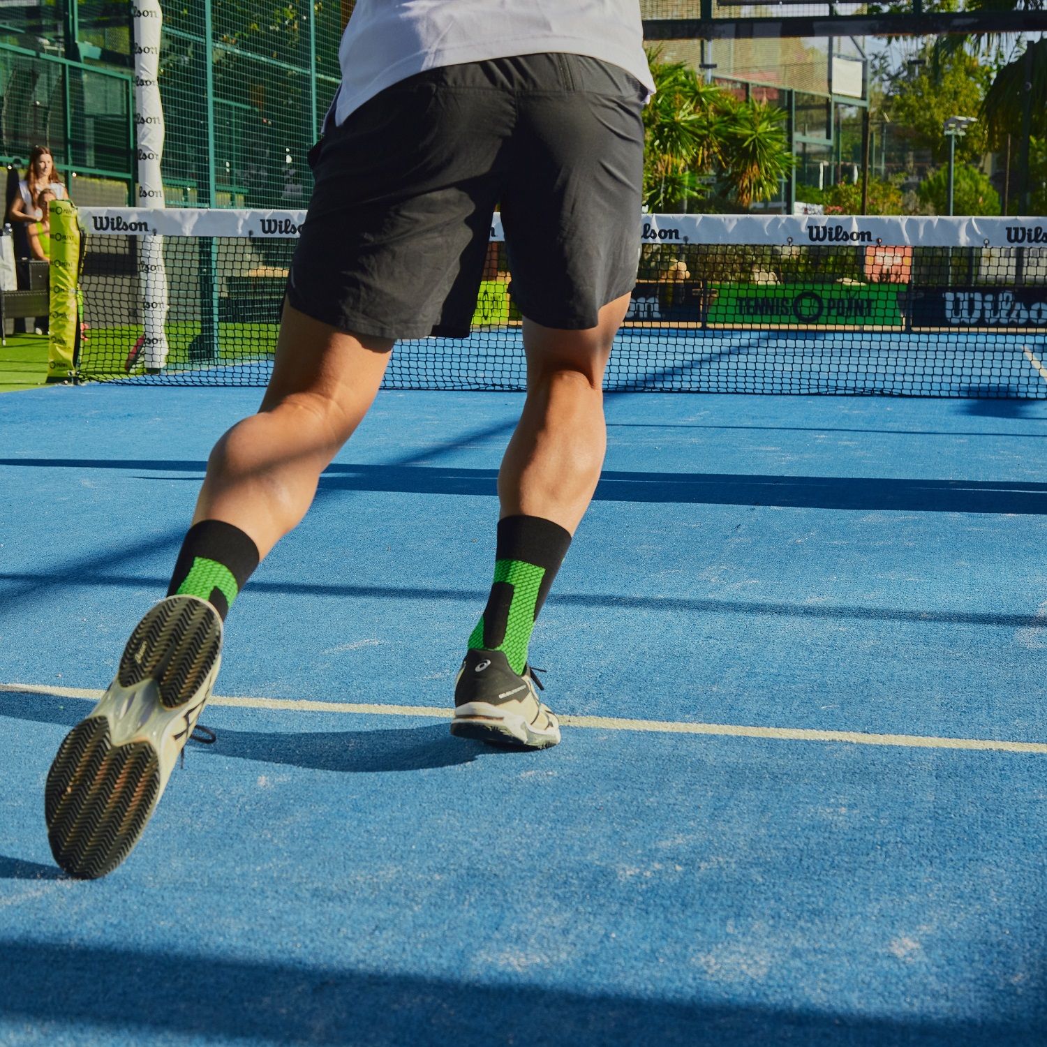 Una persona che corre su un campo da tennis indossa un calzino stabilizzatore di caviglia Enforma. I calzini sono neri con dettagli verdi. Lo sfondo mostra una rete da tennis e un campo blu.