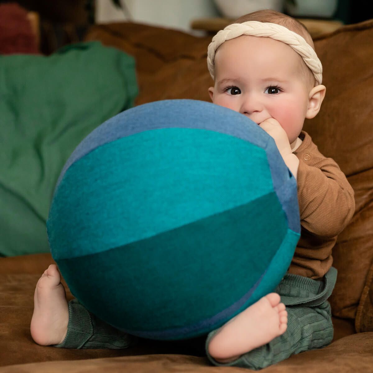 Baby sitzt mit einer blauen und grünen Stoffkugel. Das Kind hat einen Stirnband und steckt die Kugel in den Mund.