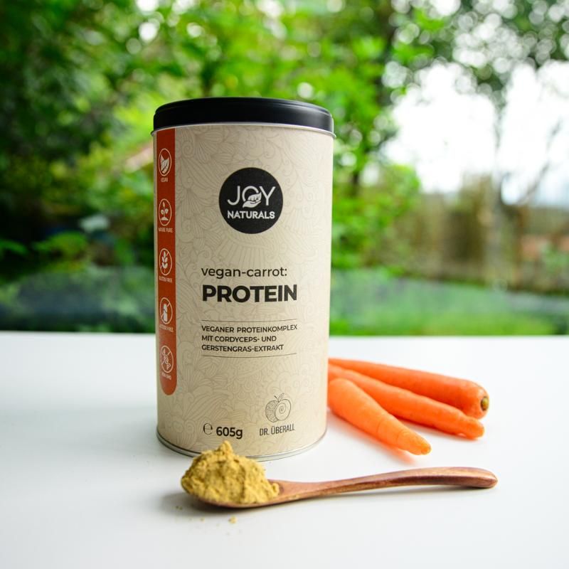 Dose neben Karotten und Pulver auf Löffel. Aufschrift: JOY NATURALS, vegan-carrot:PROTEIN, 605g. Veganer Proteinkomplex mit Cordyceps-Extrakt.
