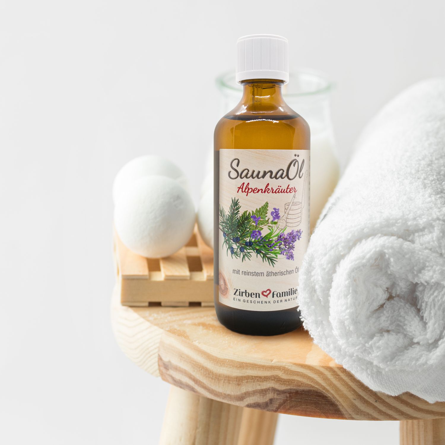 Braune Flasche mit weißem Deckel. Auf einem Holzschemel mit Handtuch und Sauna-Zubehör. Etikett mit Produktname und Illustration.