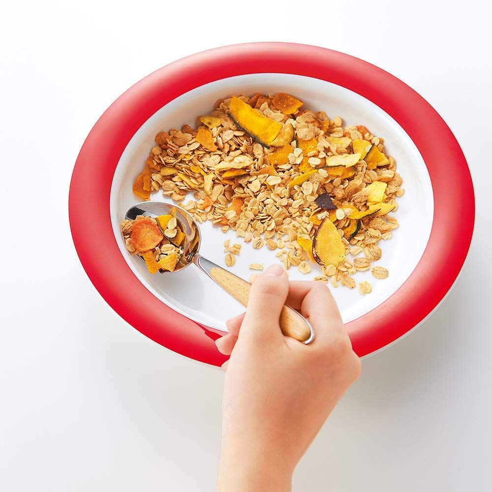 Teller mit rotem Rand, gefüllt mit Müsli und Früchten. Hand mit Löffel.