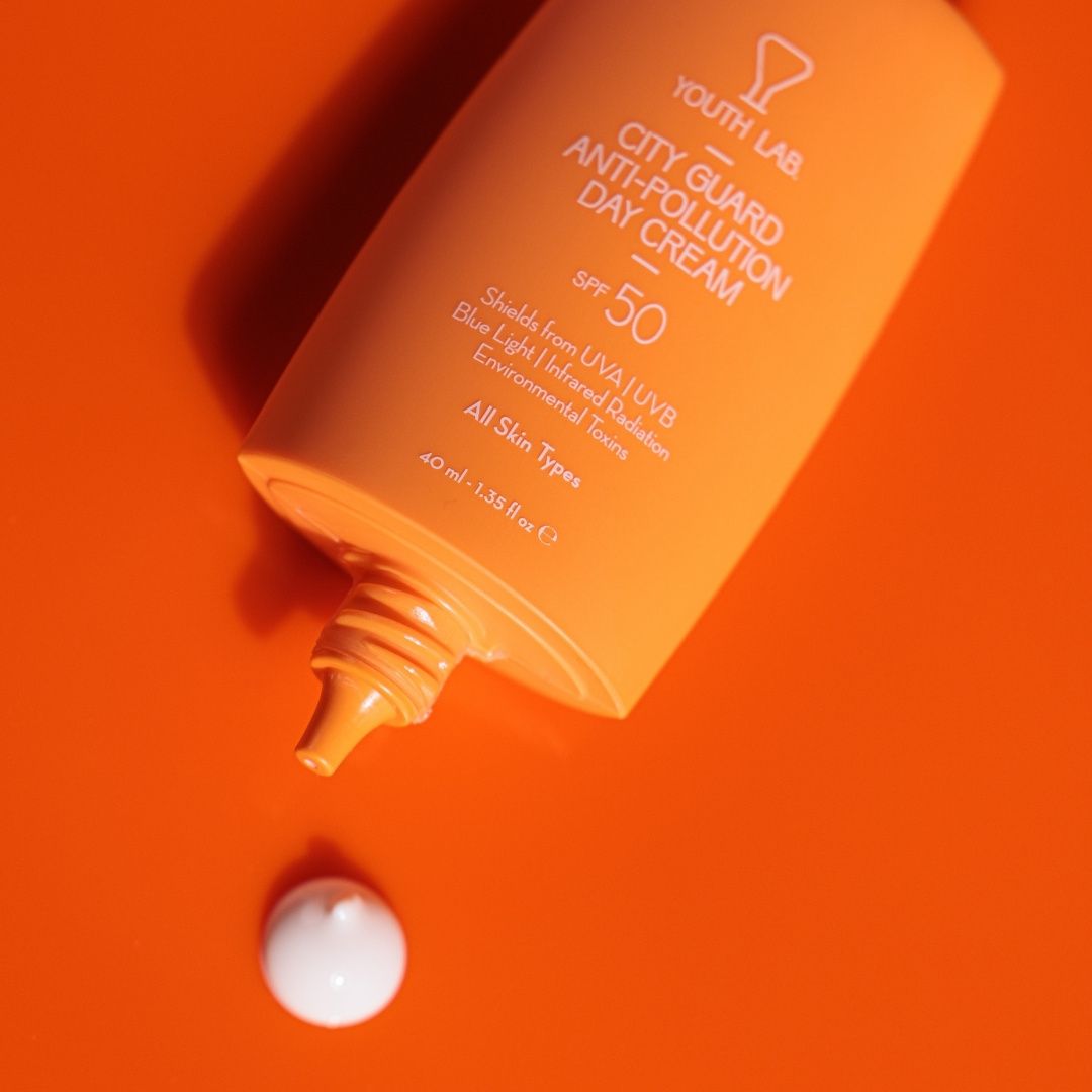 Orangefarbene Tube liegt auf orangefarbenem Untergrund. Text: YOUTH LAB, City Guard Anti-Pollution Day Cream SPF50. Ein Tropfen Creme daneben.