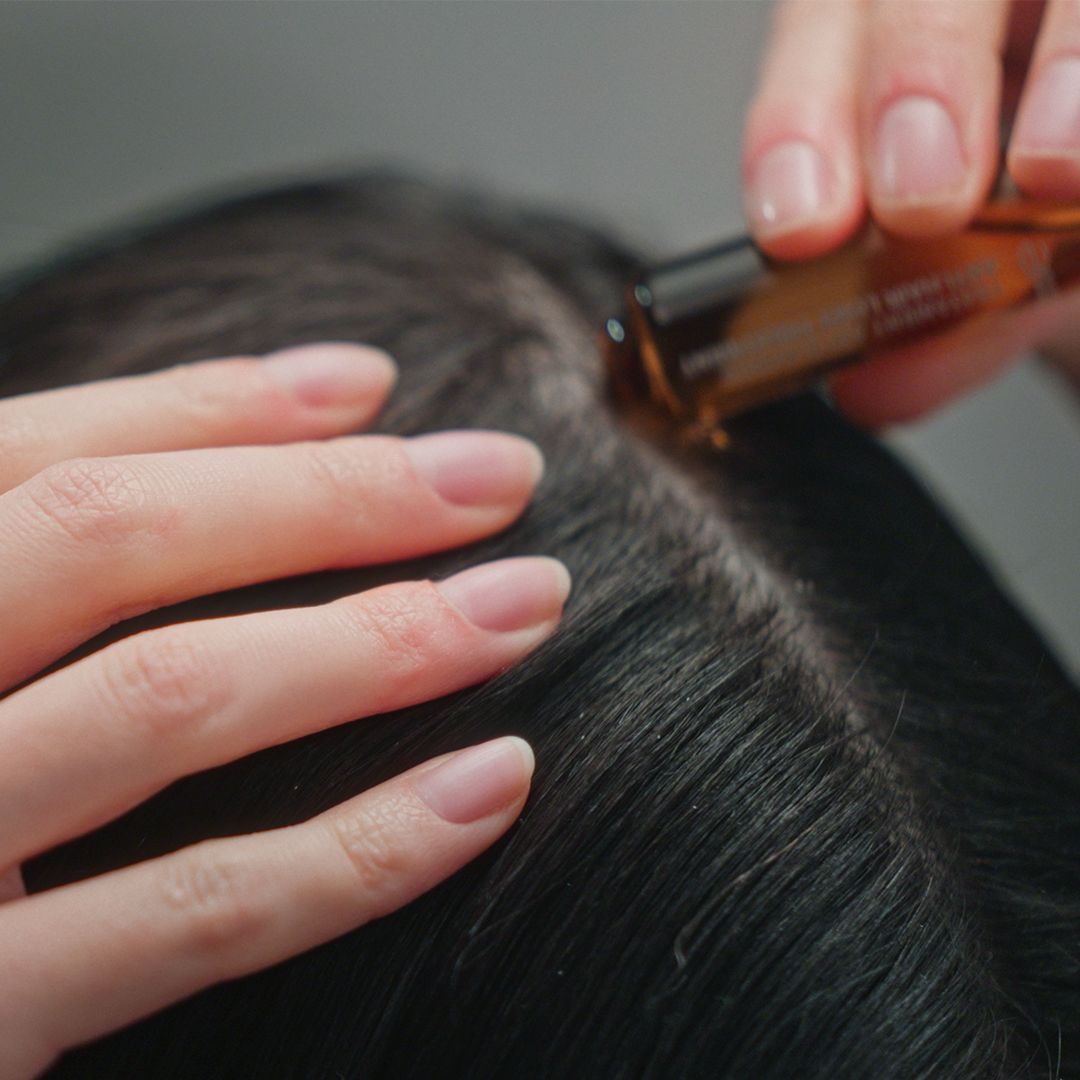 Hände tragen eine Ampulle mit brauner Flüssigkeit auf die Kopfhaut auf.