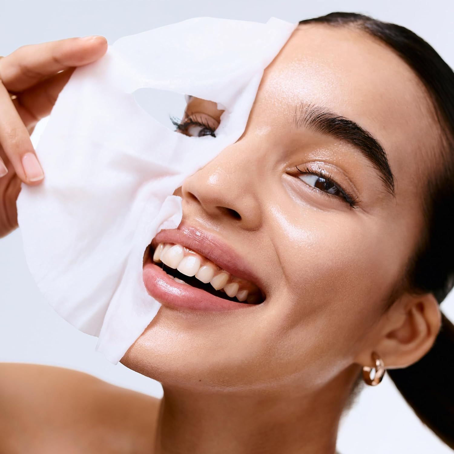 Frau hält Tuchmaske vor Gesicht. Haut glänzend, Lippen glänzend, goldener Ohrring. Maske weiß.