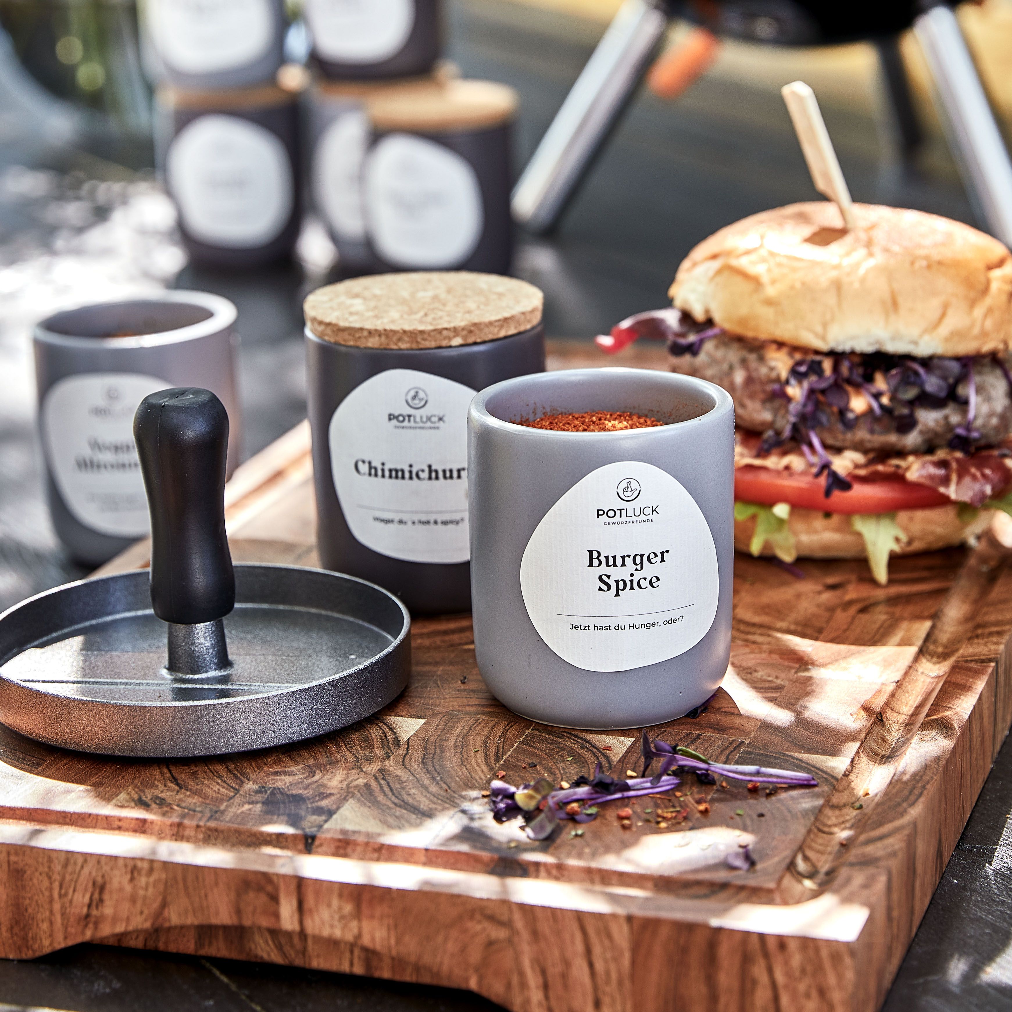 Graue Keramikbehälter mit Etiketten. Einer enthält "Burger Spice". Auf einem Holzbrett liegt ein Burger.