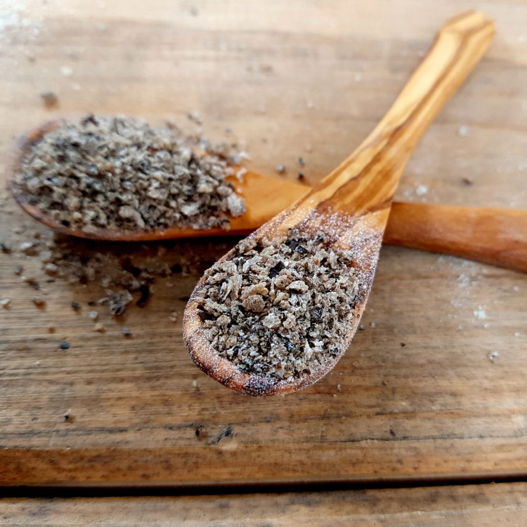 Zwei Holzlöffel mit QCHEFS Zahnputzflocken auf Holzuntergrund. Die Flocken sind grau-braun und körnig.