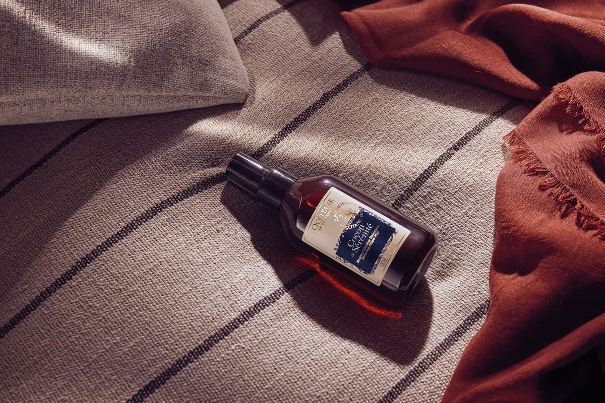 Braune Flasche mit schwarzem Sprühkopf liegt auf einer Decke. Etikett mit Produktnamen. L'Occitane Aromachologie Kopfkissen-Nebel Entspannend.