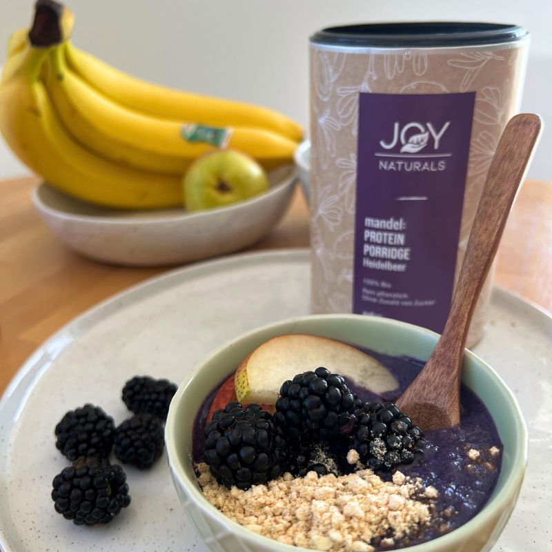 Porridge mit Blaubeeren, Mandeln, Apfel und Brombeeren in einer Schüssel. Daneben eine Dose JOY NATURALS Bio Heidelbeer Mandelprotein-Porridge.