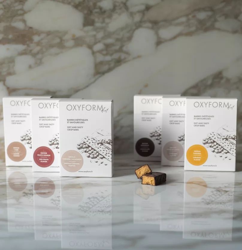 Mehrere weiße Kartons mit Oxyform-Logo. Einige Kartons stehen, andere liegen. Zwei Riegel sind angeschnitten.