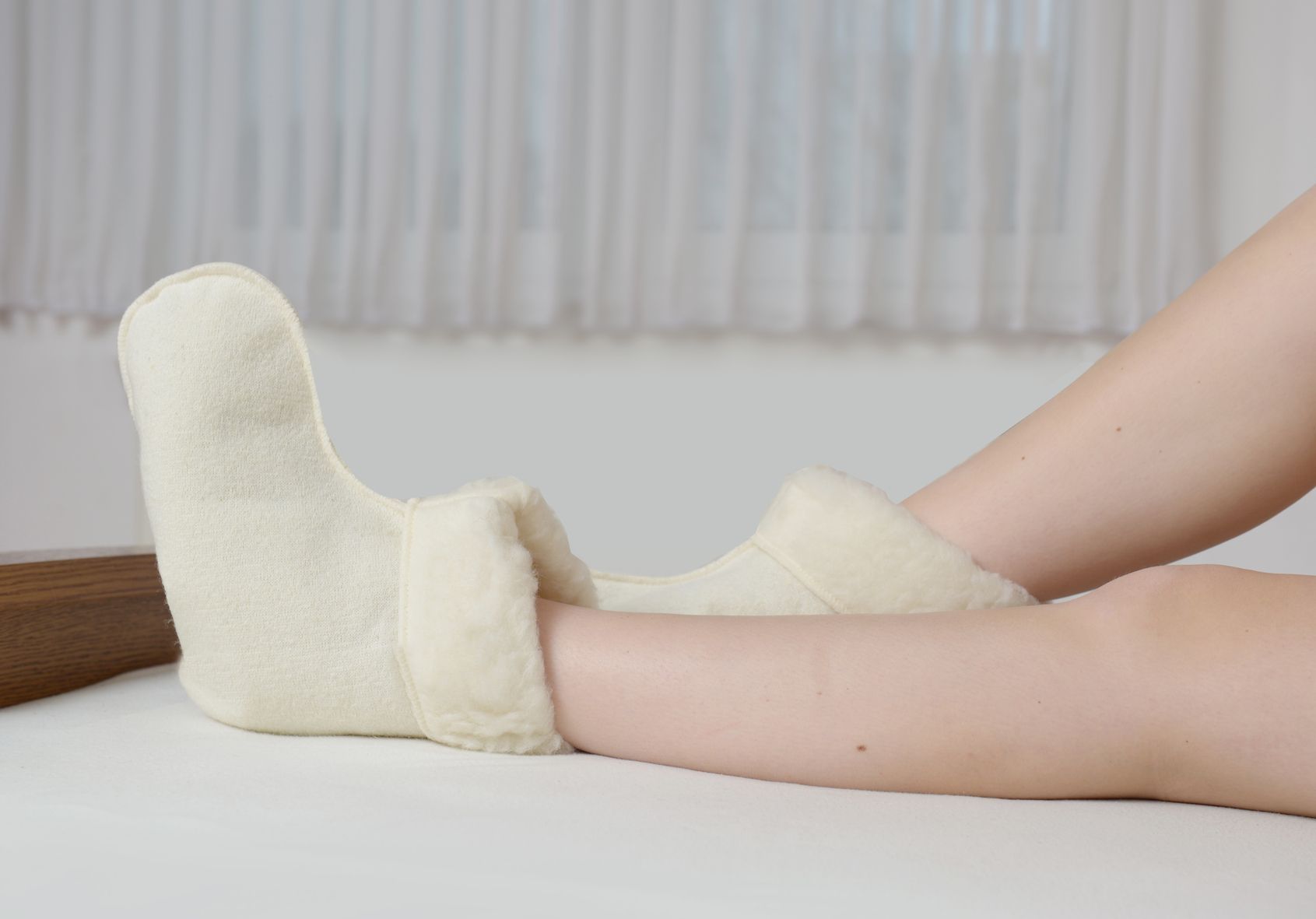 Person trägt cremefarbene Schurwoll-Bettschuhe. Füße und Unterschenkel auf weißer Unterlage.