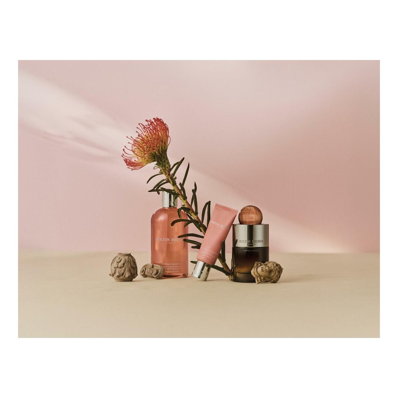Produkt-Arrangement mit Flaschen, Tube, Blütenzweig und Dekoration auf beigem Untergrund vor rosa Hintergrund.