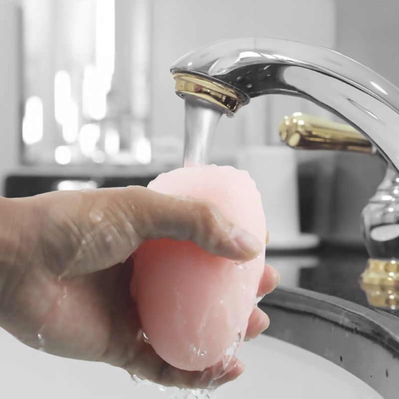Eine Hand hält ein rosa, ovales Produkt unter fließendem Wasser. Der Hintergrund ist unscharf.
