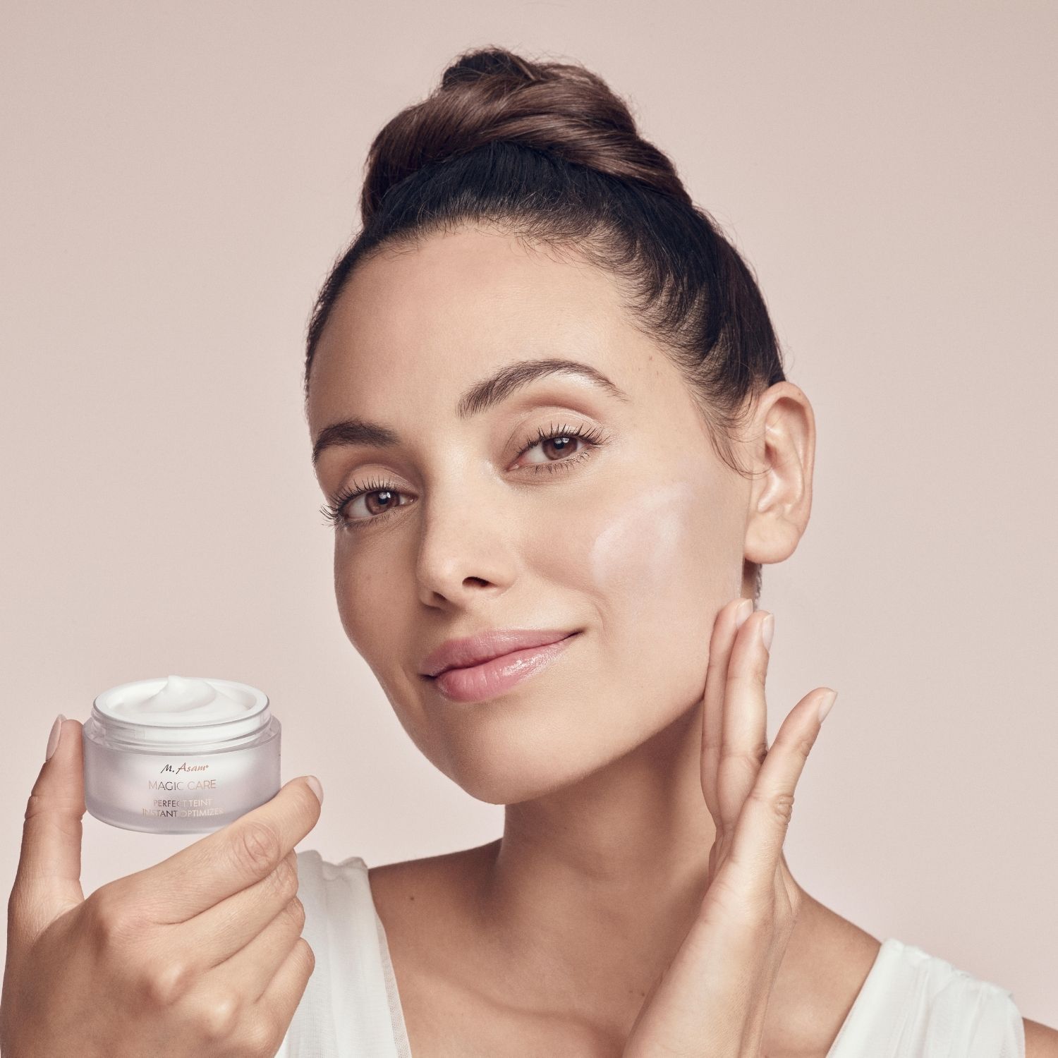 Donna che tiene un vasetto di crema. Applica la crema. Testo: M. Asam MAGIC CARE PERFECT TEINT INSTANT OPTIMIZER.