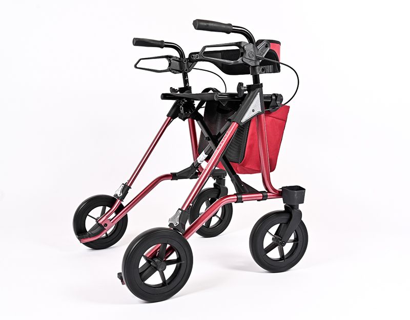 Rot-schwarzer Rollator mit vier Rädern. Er hat einen Korb, eine Rückenlehne und Handgriffe. Der Rahmen ist rot.