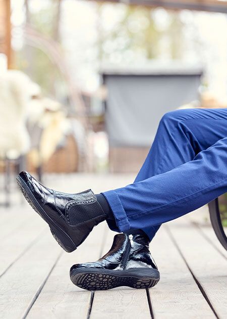 Person trägt schwarze Stiefeletten mit Reißverschluss. Lackleder-Optik, breite Passform. Auf Holzterrasse, blaue Hose.
