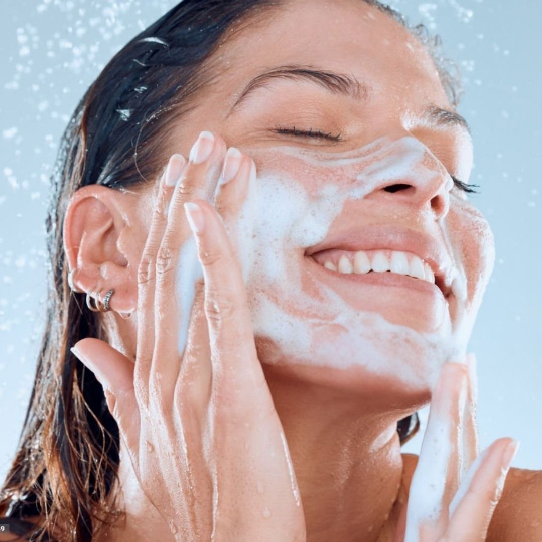 Donna che si lava il viso con la schiuma. Sorride e ha gli occhi chiusi. L'acqua le schizza sul viso.