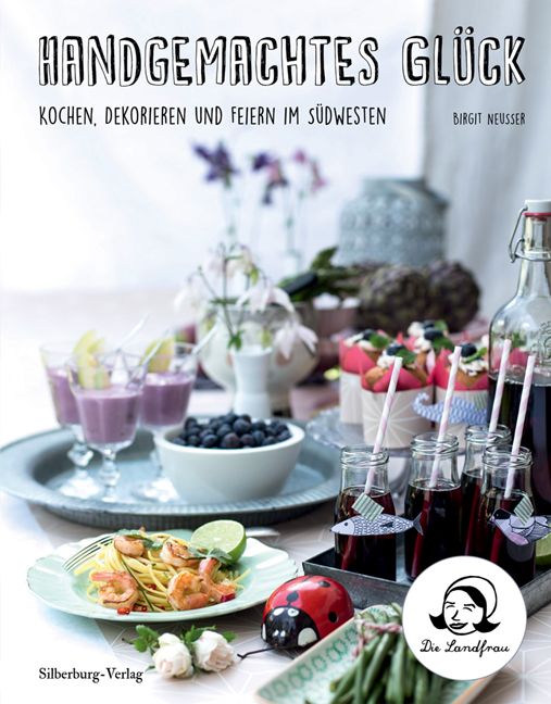 Buchcover mit Titel "Handgemachtes Glück". Darunter Text und Autor. Im Vordergrund Speisen und Getränke auf Tabletts. Logo "Die Landfrau".