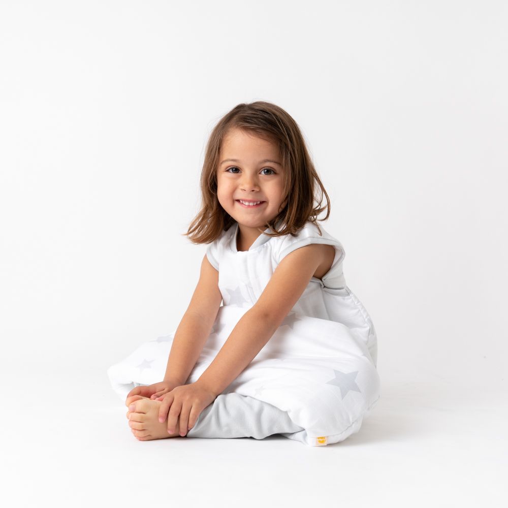 Kleines Mädchen sitzt in einem Baby-Schlafsack mit grauen Sternen. Lächelt in die Kamera.