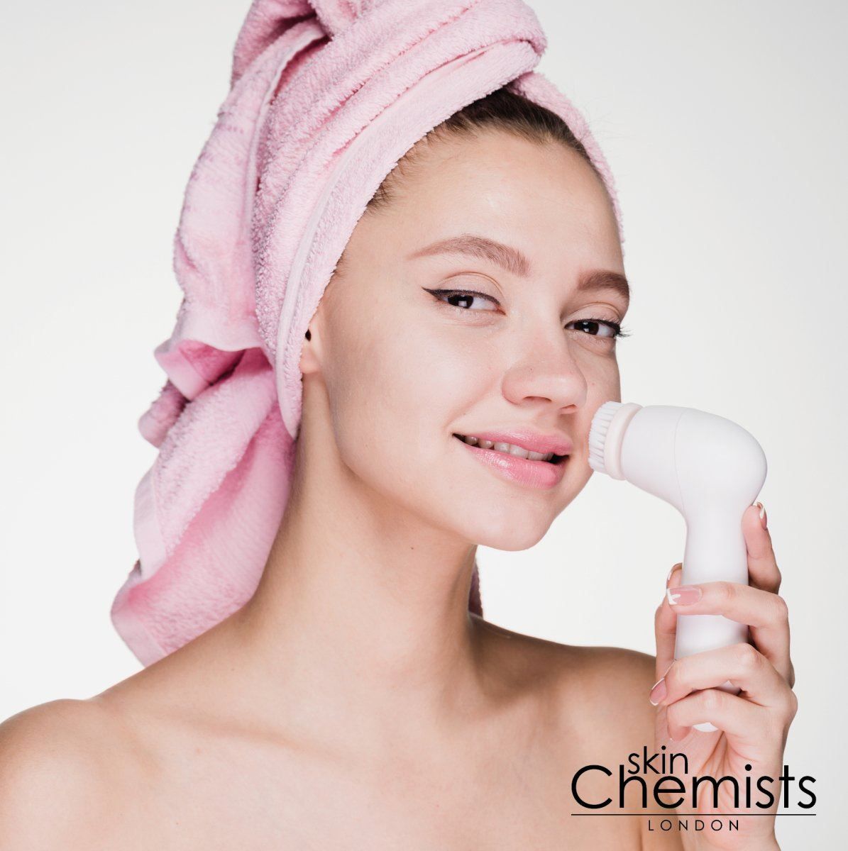 Frau mit rosa Handtuch benutzt weiße Gesichtsreinigungsbürste. Skin Chemists Logo.