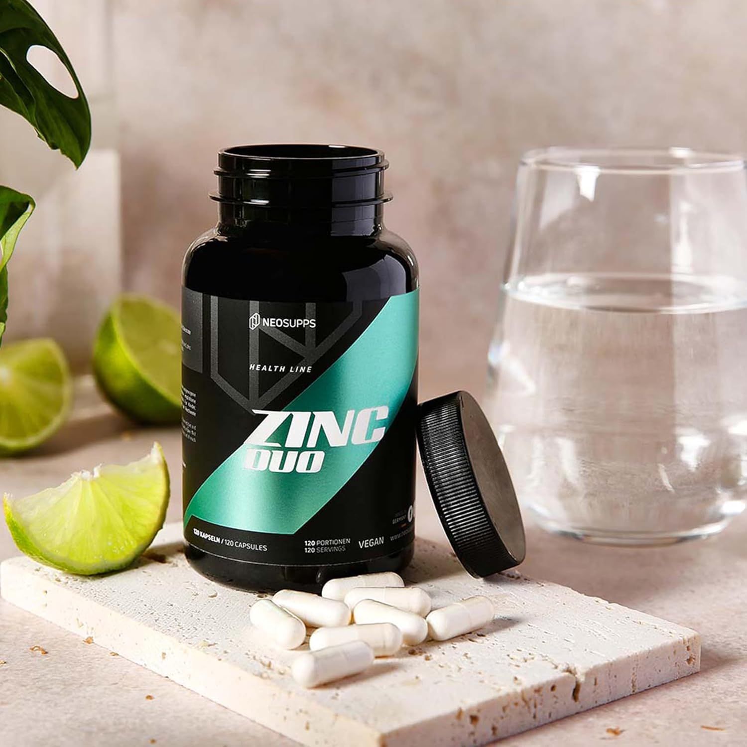Schwarze Flasche "ZINC DUO" mit geöffnetem Deckel. Daneben Kapseln und ein Glas Wasser. Limettenscheiben auf einem Tablett.