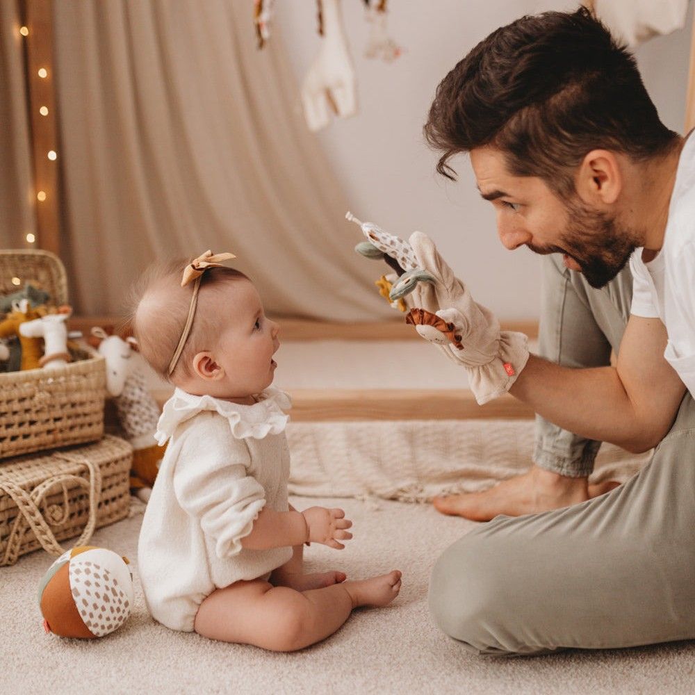 Vater spielt mit Baby. Fingerspielfigur Giraffe, beige, mit Giraffen-, Löwen- und Blattformen. Baby sitzt auf Teppich.