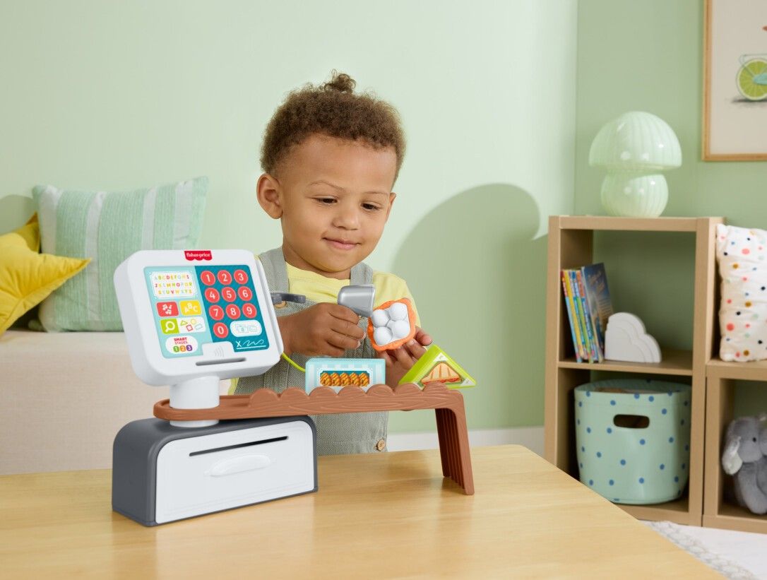 Kind spielt mit Spielzeugkasse. Kasse mit Zubehör auf Tisch. Fisher-Price Logo sichtbar.