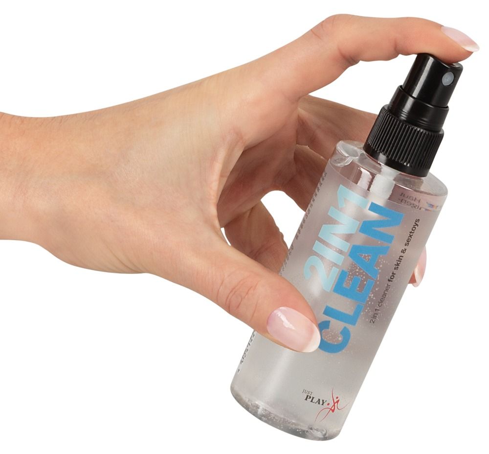 Hand hält eine transparente Flasche mit Sprühkopf. Aufschrift "2IN1 CLEAN" und "2in1 cleaner for skin & sextoys". Marke: Just Play.