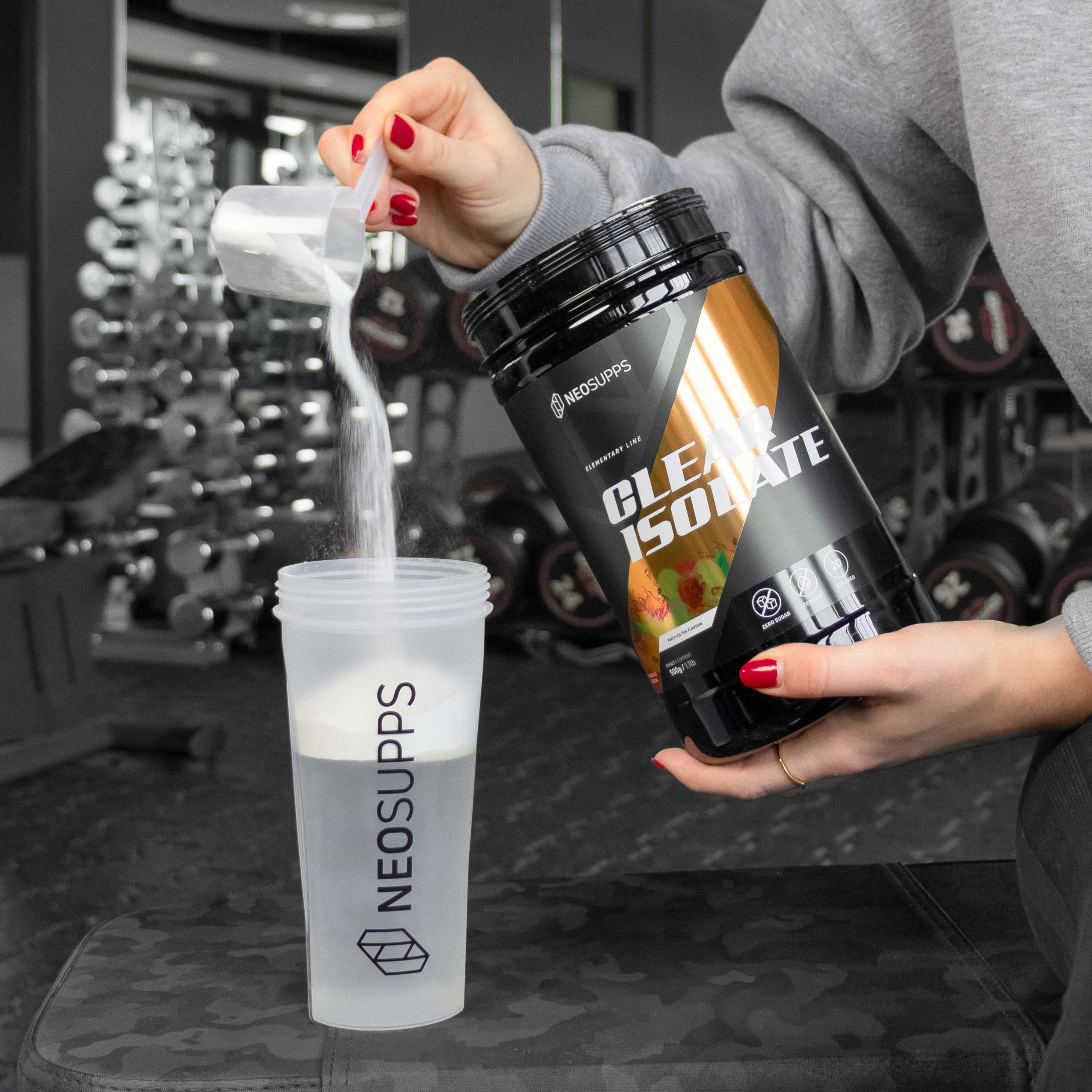 Person gießt Pulver aus Dose "CLEAR ISOLATE" in Shaker. Marke: NEOSUPPS. Fitnessstudio-Hintergrund.