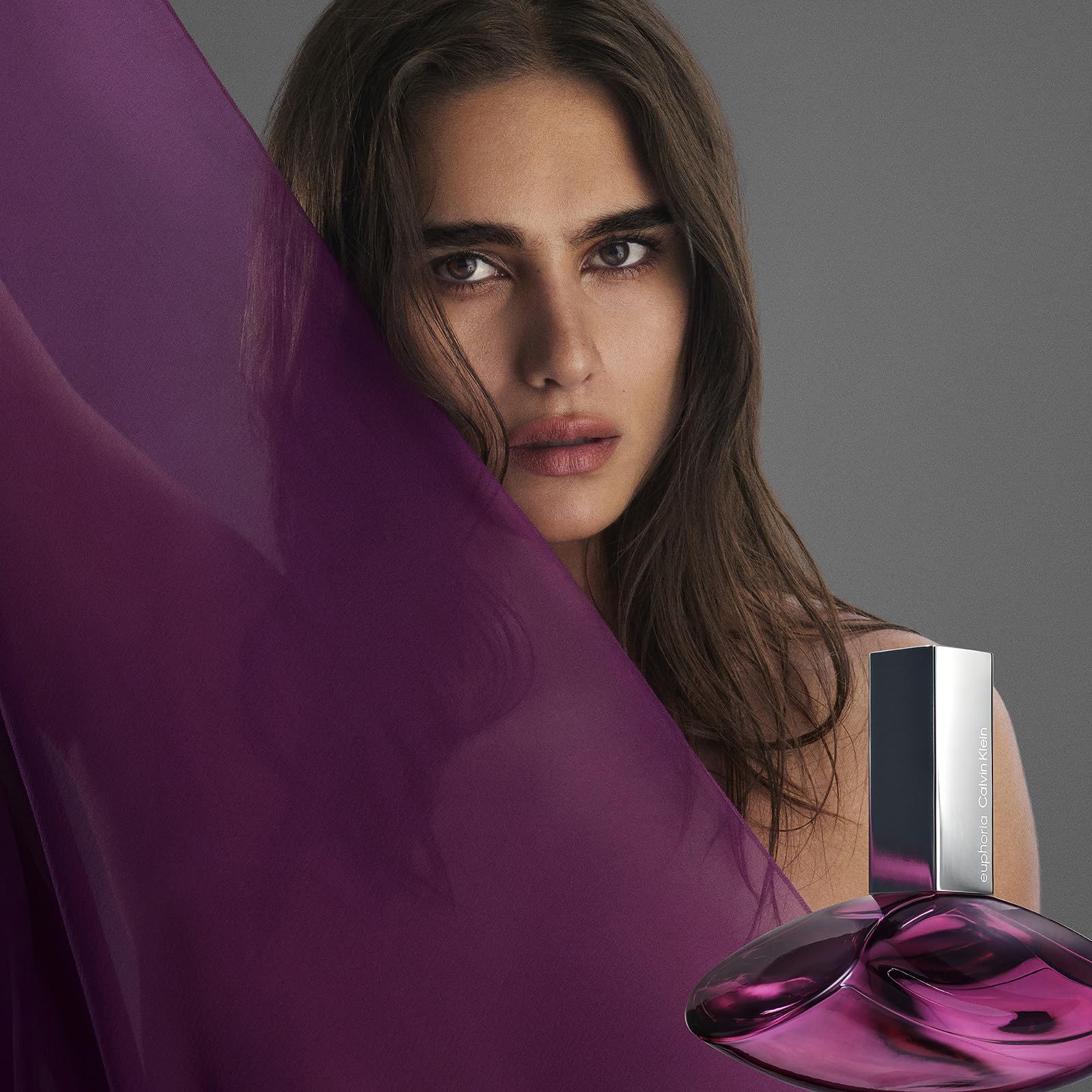 Frau mit Parfümflasche. Flasche silberfarben, tropfenförmig. Frau hält violetten Stoff.