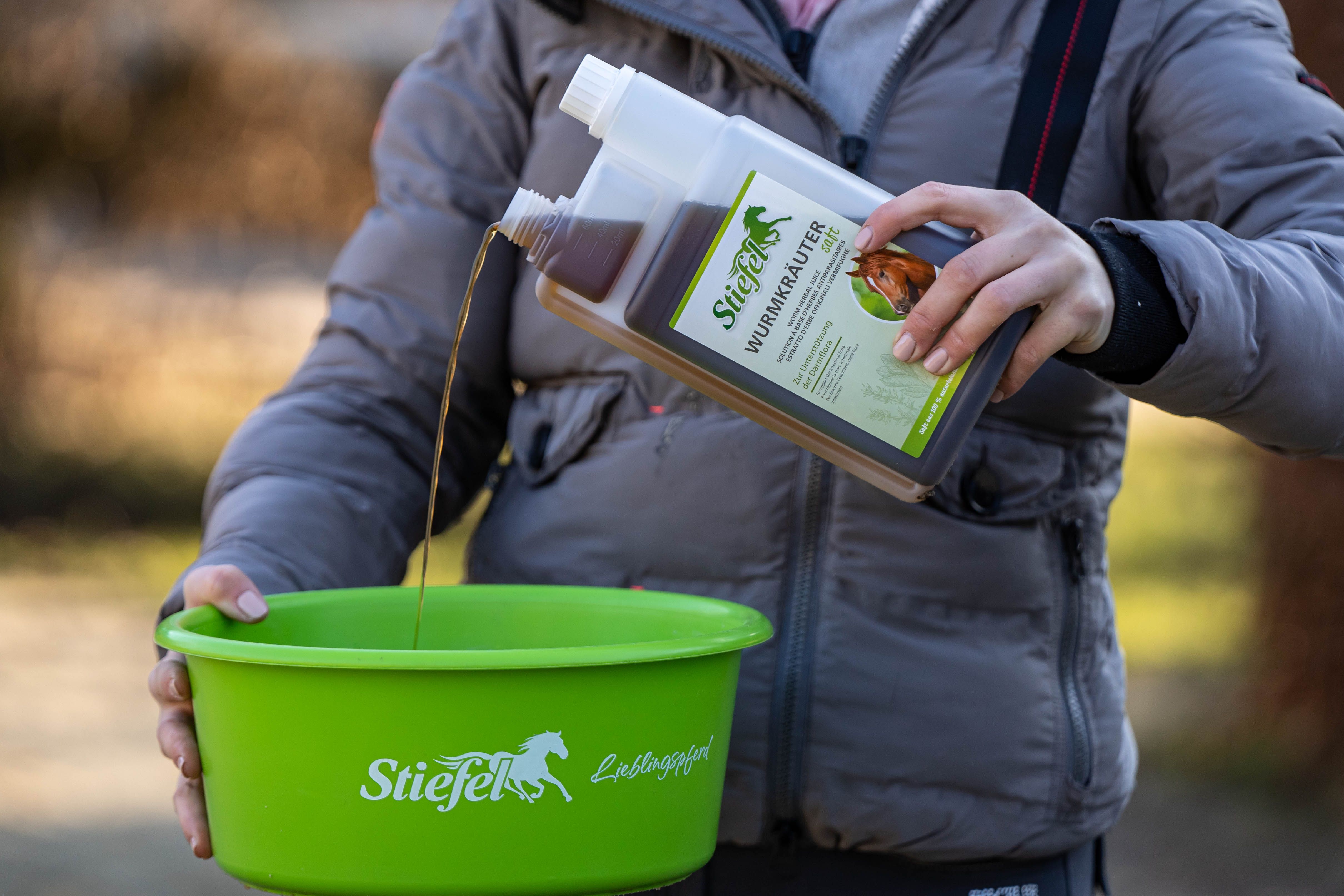 Person gießt braune Flüssigkeit aus einer Doppelkammerflasche in einen grünen Behälter. Aufschrift: Stiefel Wurmkräuter.
