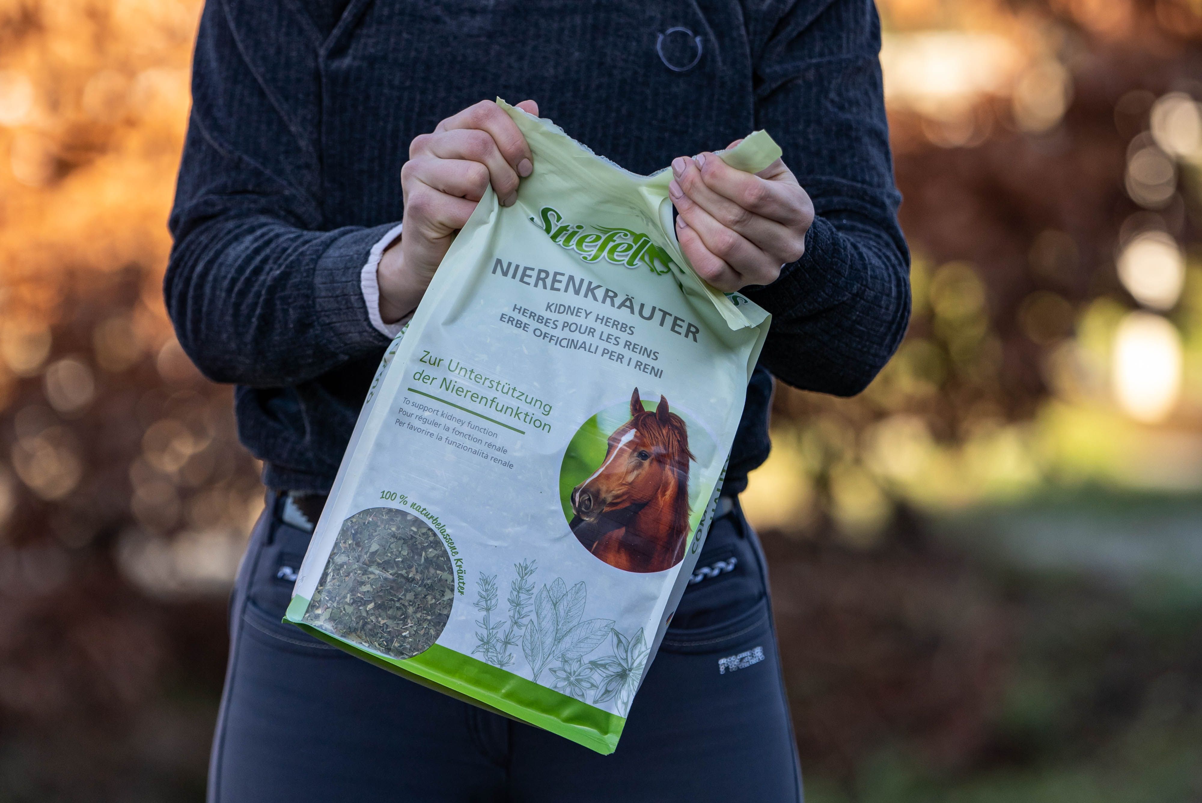 Person hält einen Beutel Stiefel Nierenkräuter. Aufschrift: Kidney Herbs. Abbildung eines Pferdes. Hintergrund: verschwommene Natur.