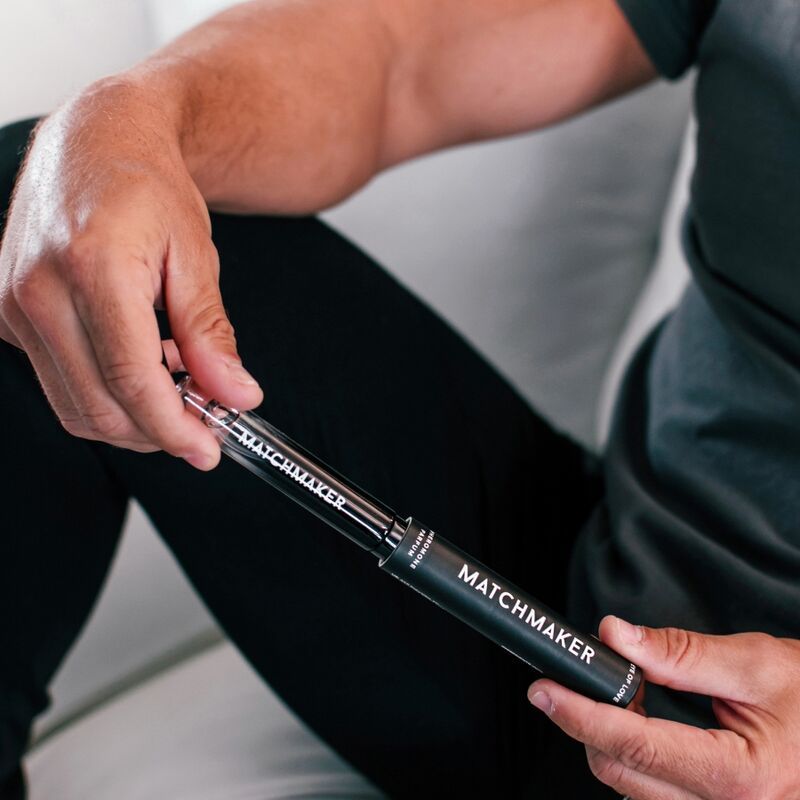 Mann hält die Verpackung und den Flakon. Schwarze zylindrische Verpackung und Flakon mit weißem Schriftzug MATCHMAKER.