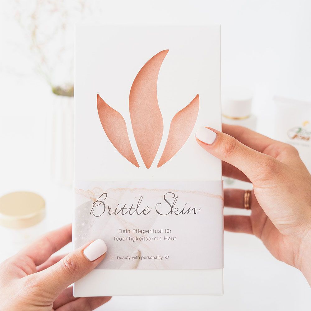 Hände halten eine weiße Verpackung mit einem ausgeschnittenen Logo. Auf der Verpackung steht 'Brittle Skin' und weitere Texte. Produkte im Hintergrund.