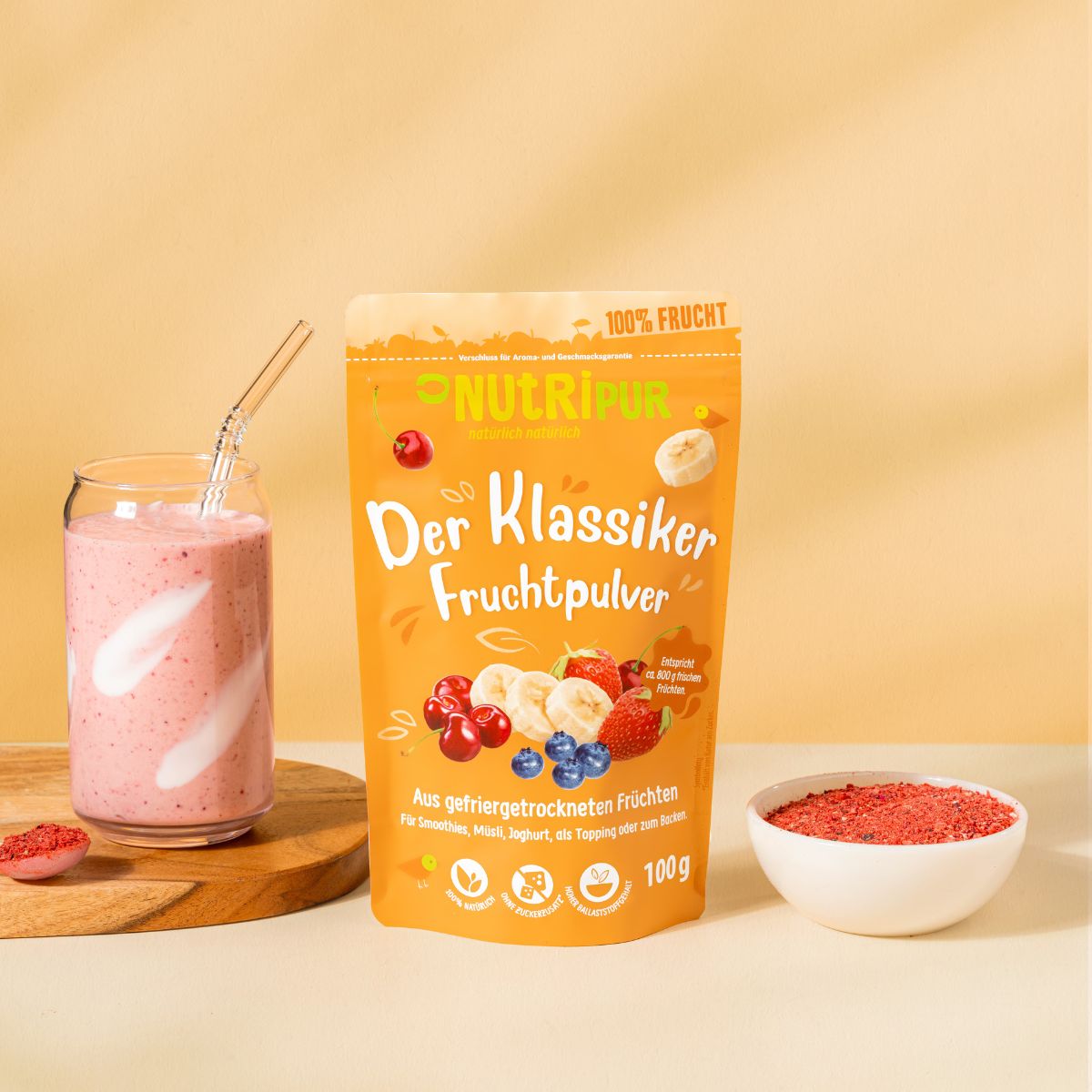 Glas mit rosa Smoothie, Strohhalm. Daneben eine Tüte mit Fruchtpulver und eine Schale mit Fruchtpulver. Auf einem Holzbrett.