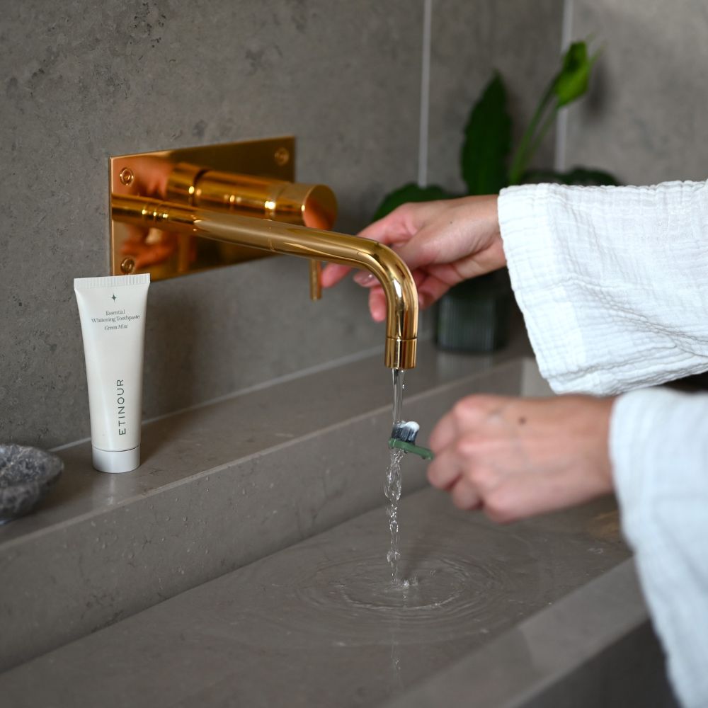 Zahnpasta-Tube neben Waschbecken. Person wäscht Zahnbürste unter Wasserhahn. Zahnpasta-Tube mit Text: Essential Whitening Zahnpasta.